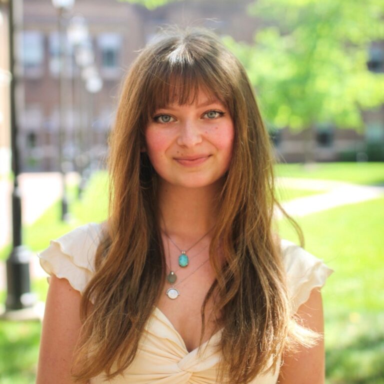 Ella Balzano, a young woman with long brown hair, wears a cream-colored top and necklaces.