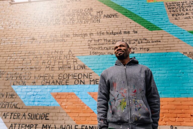Man smiles in front of a brick mural with inspirational text