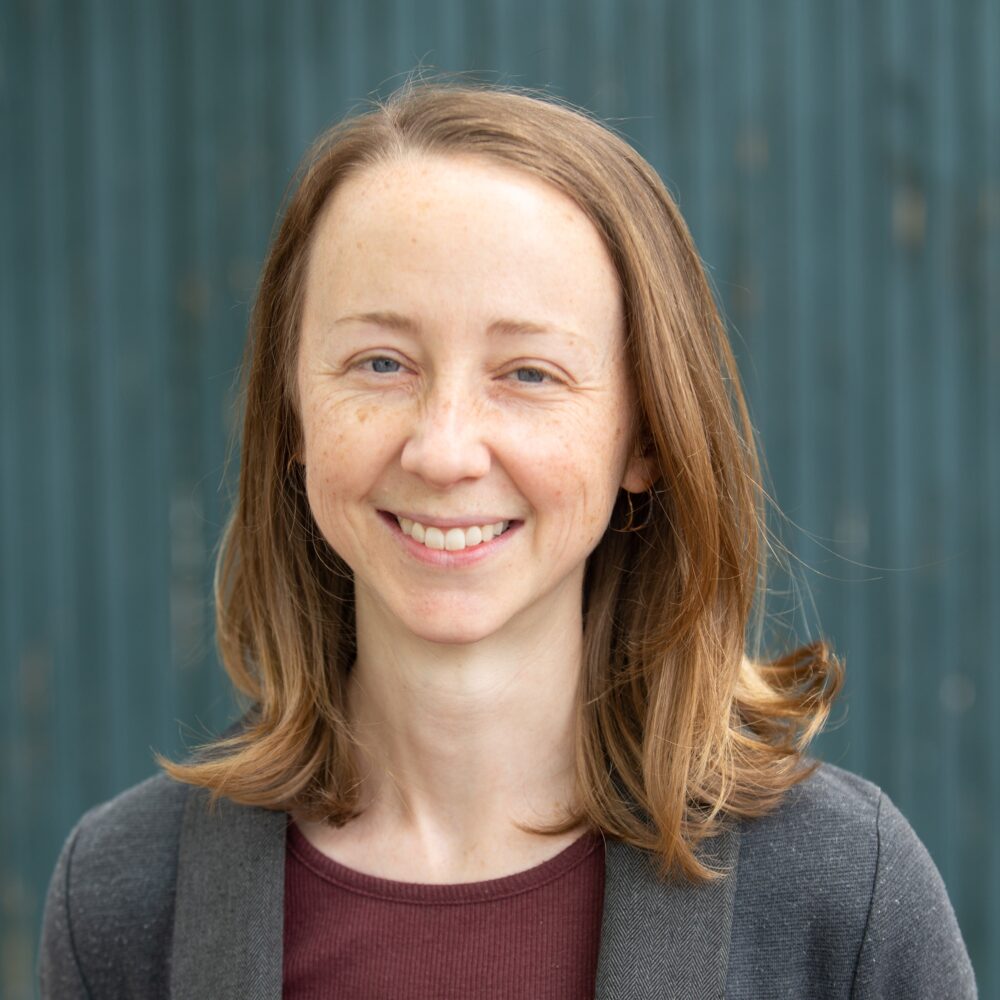 Hannah Ayers smiling, with freckles and blue eyes, wearing a grey jacket.