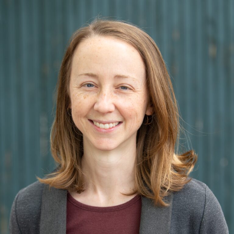 Hannah Ayers smiling, with freckles and blue eyes, wearing a grey jacket.