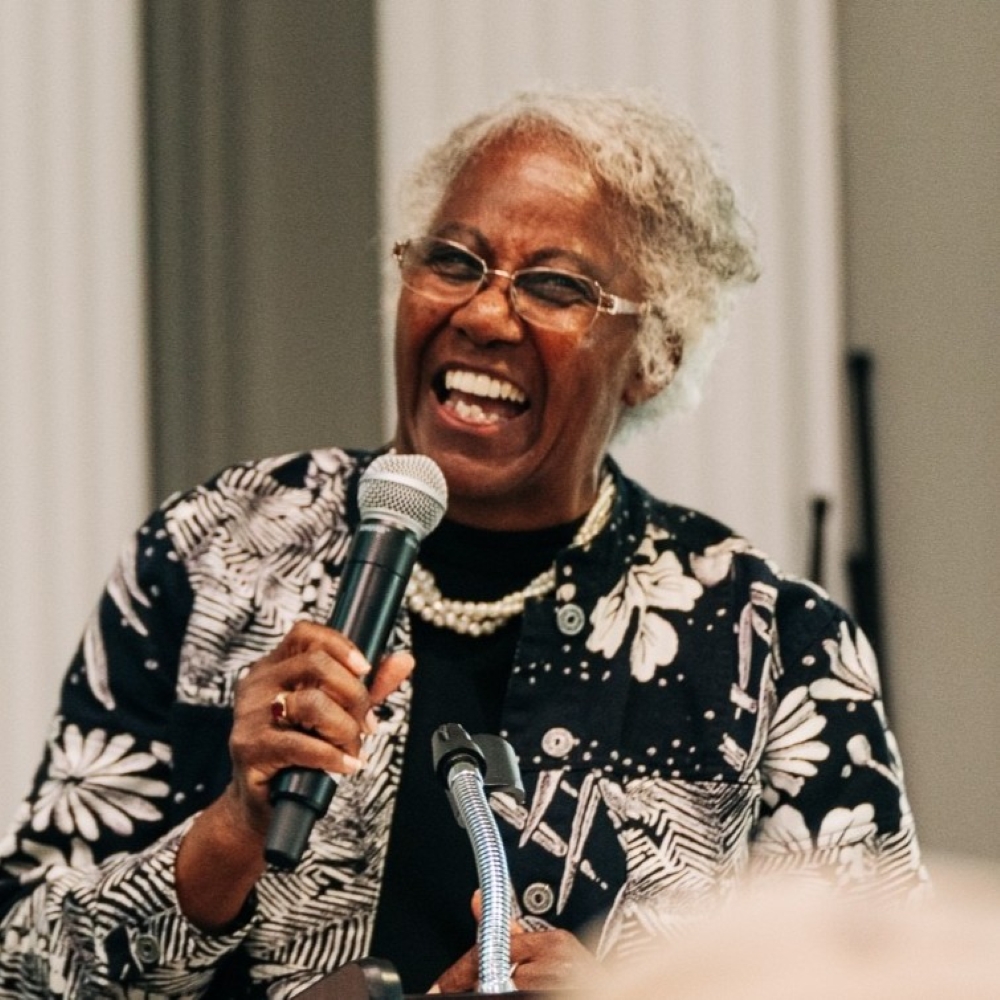 Helena Estes-Johnson speaks animatedly into a microphone, wearing a patterned jacket.