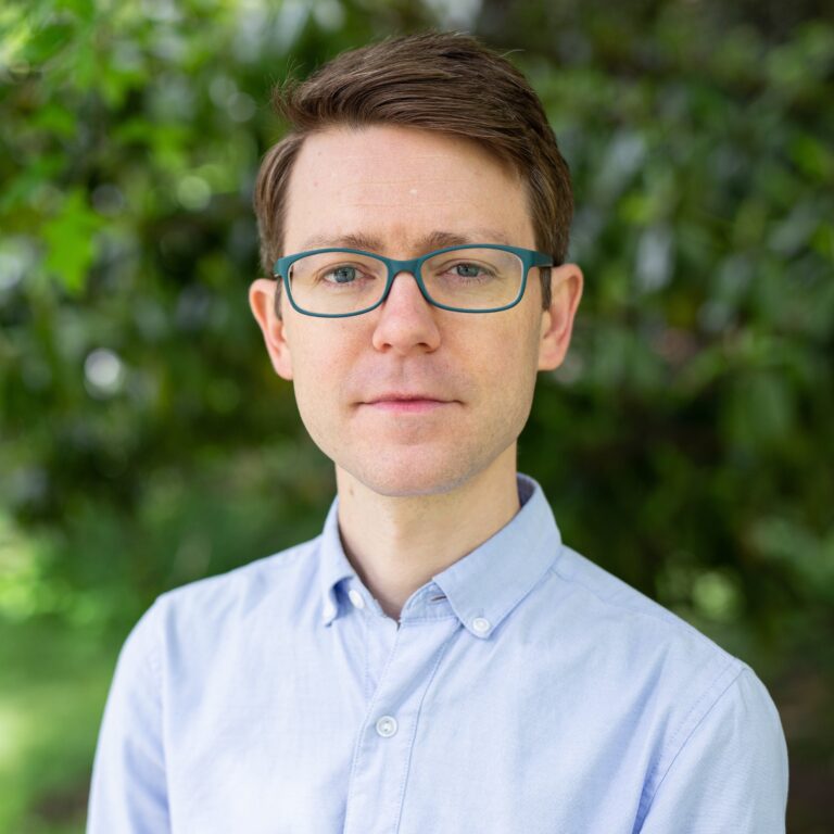 Lance Warren, a man with glasses, wears a blue shirt outdoors.